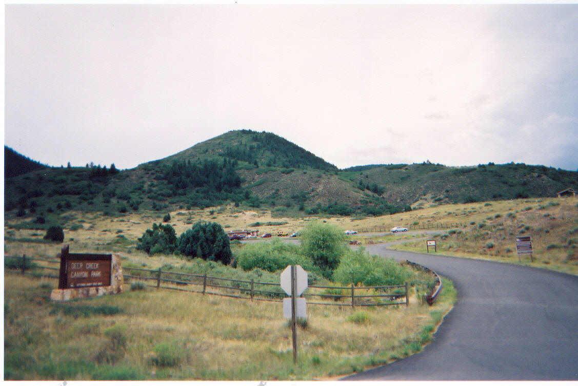 A winding road leading into a canyon park, surrounded by rolling hills and greenery. A sign for "Deep Creek Canyon Park" is prominently displayed on the left, and a few parked cars are visible in the background. The scene is set under a cloudy sky, indicating an overcast day. Deer Creek Canyon mountain bike trail.