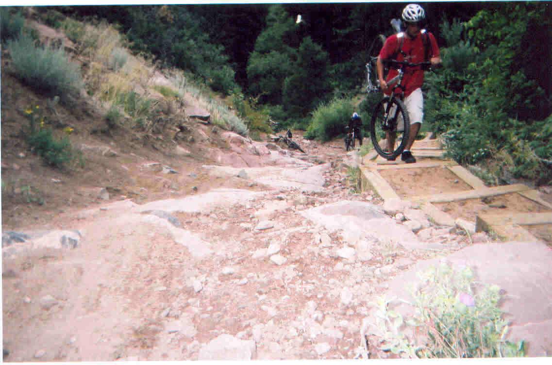 A mountain biker pushes their bike uphill on a rocky, uneven path surrounded by greenery. The narrow trail features wooden beam sections to aid navigation, with another biker visible further up the trail. Deer Creek Canyon mountain bike trail.