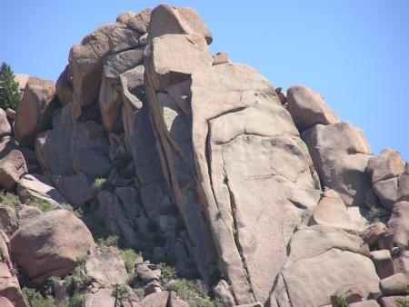 A large rock formation, with prominent cliffs and smooth surfaces, surrounded by smaller boulders and vegetation against a clear blue sky. Monument Preserve mountain bike trail.