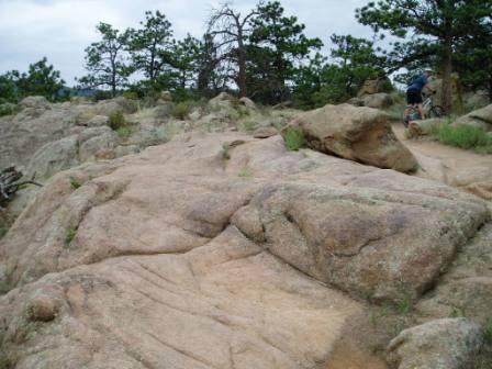 A rugged rocky landscape with a cyclist riding along a path. The scene features large boulders and scattered vegetation, with pine trees in the background under a cloudy sky. Hall Ranch mountain bike trail.