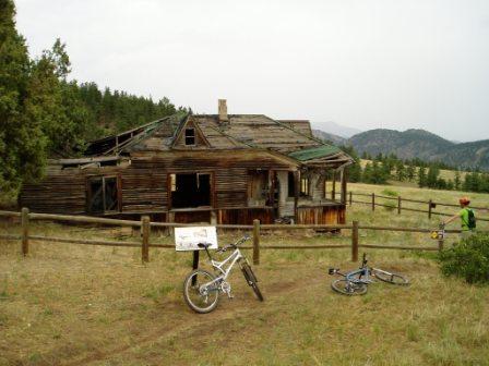 An old, abandoned house with a deteriorating wooden structure, surrounded by a grassy field and mountains in the background. Two bicycles are parked nearby, and a sign stands in front of the house, indicating it as a point of interest. The scene is set in a natural, outdoor environment with trees and hills visible. Hall Ranch mountain bike trail.