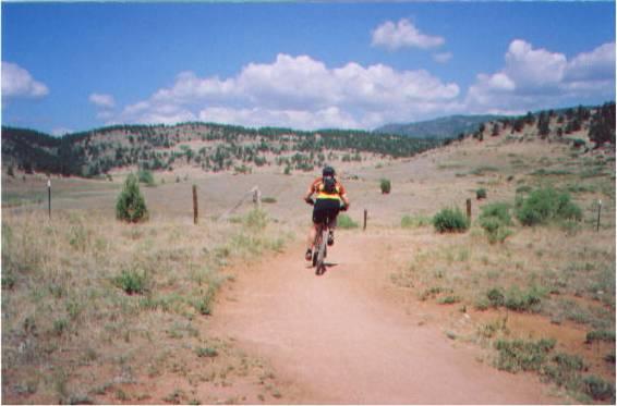 A person riding a bike on a dirt path through a grassy landscape, with rolling hills and a blue sky filled with clouds in the background. Hall Ranch mountain bike trail.