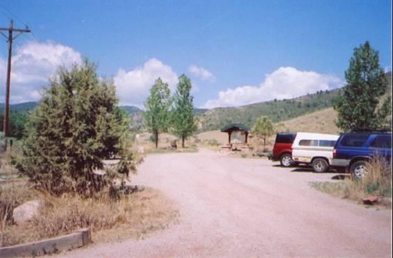 A gravel parking area surrounded by trees and mountains under a clear blue sky, featuring several parked vehicles and a small covered shelter in the background. Hall Ranch mountain bike trail.