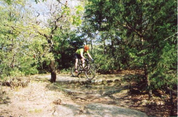 A mountain biker in a bright green shirt and orange helmet performs a jump on a rocky trail surrounded by trees. Sunlight filters through the foliage, creating a vibrant outdoor scene. McMurtry Trail mountain bike trail.