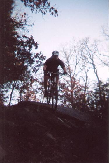 A mountain biker jumping off a rock on a forest trail during sunset, surrounded by trees with autumn foliage. McMurtry Trail mountain bike trail.