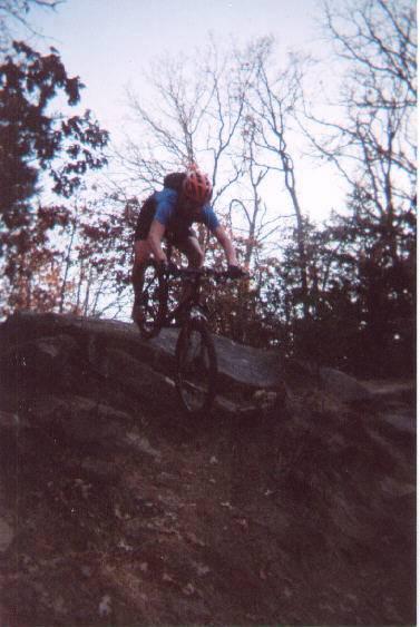 A mountain biker in a helmet jumps off a rocky ledge, surrounded by trees in a natural setting. The bike is airborne, capturing the action-packed moment. McMurtry Trail mountain bike trail.