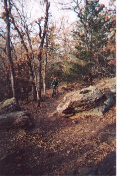 A cyclist riding along a dirt trail surrounded by trees and rocky outcrops in a forested area during autumn, with fallen leaves scattered on the ground. McMurtry Trail mountain bike trail.