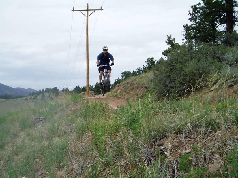 A mountain biker in a blue shirt and helmet jumps over a trail in a grassy, hilly area with trees and power lines in the background. The sky is overcast, indicating a cloudy day. Hall Ranch mountain bike trail.