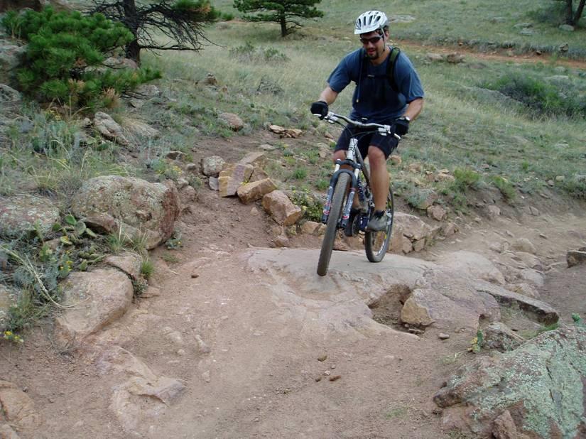 A person riding a mountain bike on a rocky trail, navigating over a section of exposed rock and dirt. The background features a grassy area with scattered rocks and trees. The cyclist is wearing a helmet and casual sports attire. Hall Ranch mountain bike trail.