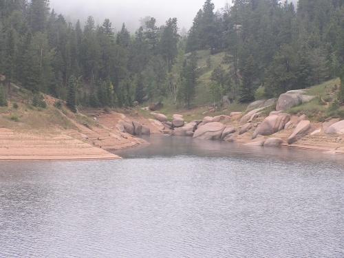 A peaceful lakeside scene featuring calm waters surrounded by rocky shores and lush green trees in the background, with a misty atmosphere. Rampart Reservoir mountain bike trail.