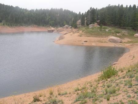A serene landscape featuring a calm lake surrounded by sandy shores and rocky outcrops, with lush green trees in the background. The scene is slightly shrouded in mist, creating a tranquil and peaceful atmosphere. Rampart Reservoir mountain bike trail.