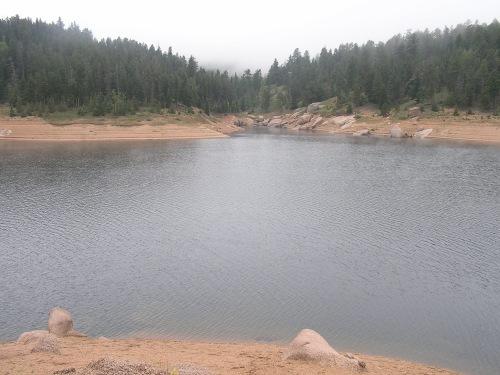 A tranquil lake surrounded by a sandy shoreline and dense evergreen trees. The water is calm and reflects the overcast sky, with fog rolling over the distant hills. Rampart Reservoir mountain bike trail.