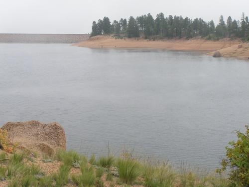Calm lake surrounded by sandy shore and pine trees under a cloudy sky. The water has a slight mist, reflecting the tranquility of the landscape. Rampart Reservoir mountain bike trail.