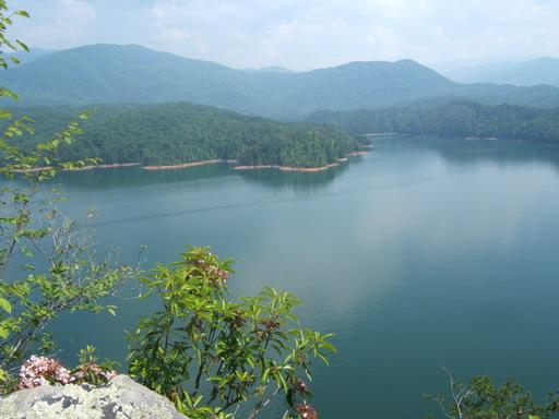 Scenic view of a tranquil lake surrounded by lush green mountains under a cloudy sky, with reflections of the landscape in the water. Tsali Right Loop mountain bike trail.