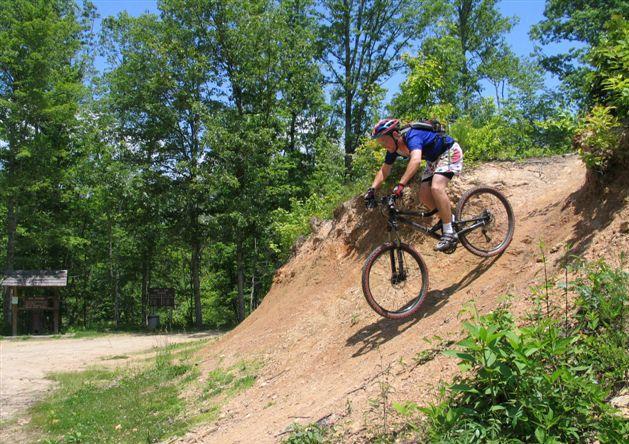 A person in a blue and white cycling outfit rides a mountain bike down a dirt slope in a wooded area, surrounded by greenery. The scene captures the thrill of mountain biking in nature. Tsali Left Loop mountain bike trail.
