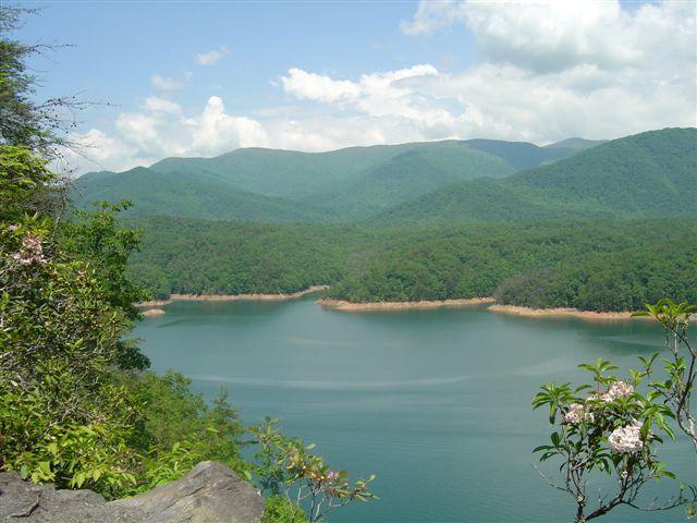 A scenic view of a tranquil lake surrounded by lush green mountains under a partly cloudy sky. The shoreline features sandy areas with trees and flowering plants, while the calm water reflects the surrounding landscape. Tsali Mouse Branch Loop mountain bike trail.