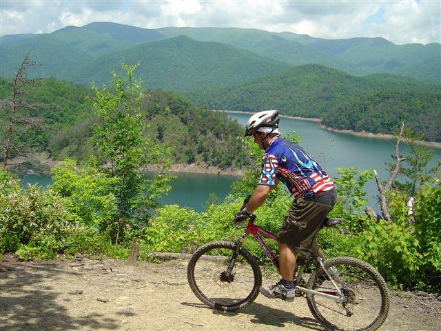 A mountain biker stops to admire the scenic view of a lake surrounded by lush green hills on a sunny day. The rider is wearing a colorful cycling jersey and helmet, with trees and rocky terrain nearby. Tsali Mouse Branch Loop mountain bike trail.