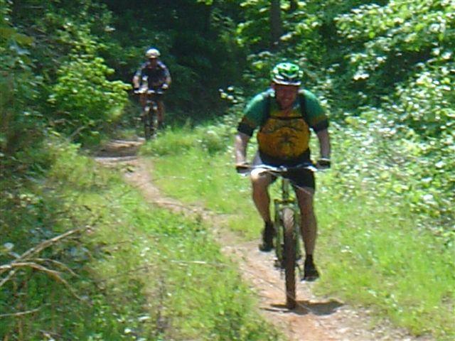 Two cyclists riding on a dirt trail surrounded by greenery. The cyclist in the foreground is wearing a green helmet and a yellow and green jersey, while the cyclist in the background is in a dark outfit. The setting is sunny and vibrant, indicating a recreational outdoor activity in nature. Tsali Thompson Loop mountain bike trail.
