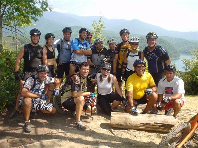 A group of fifteen mountain bikers, wearing helmets and various cycling gear, posing together outdoors near a lake with mountains in the background. They are gathered on a dirt trail, some sitting on a log, while others stand behind them, smiling at the camera. The scene captures a sunny day and a sense of camaraderie among the cyclists. Tsali Mouse Branch Loop mountain bike trail.