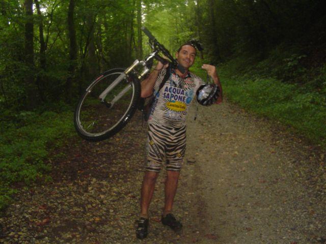 A cyclist wearing a zebra-patterned jersey and shorts is smiling and posing triumphantly while holding a mountain bike on one shoulder. The background features a lush, green forest path. The cyclist is also holding a helmet in his hand. Tsali Mouse Branch Loop mountain bike trail.