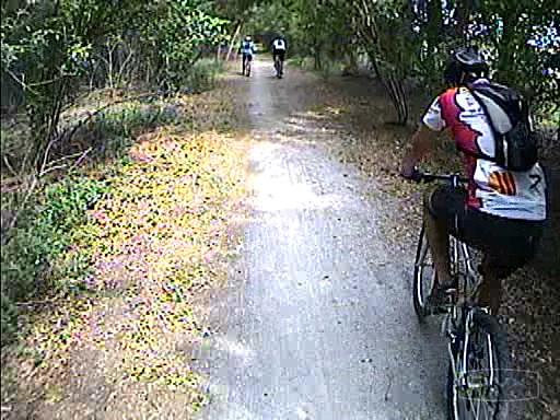 A cyclist riding on a dirt path surrounded by trees, with two other cyclists visible in the distance. The scene captures a peaceful outdoor setting, showcasing the enjoyment of biking in nature. Tom Brown / Lafayette Heritage Park mountain bike trail.