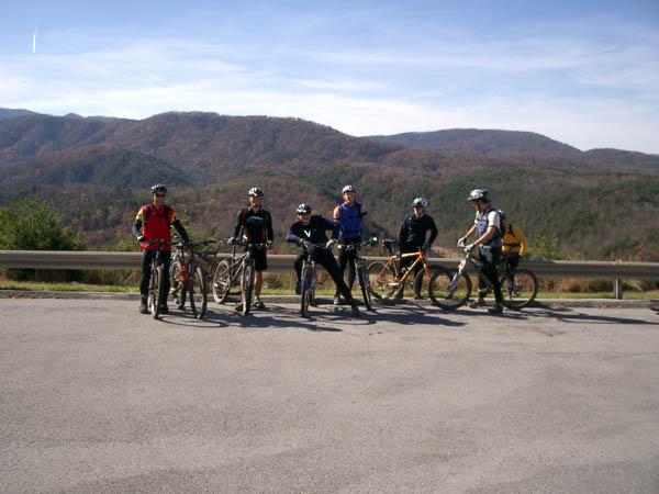 A group of six mountain bikers posing together on a scenic road with rolling hills and colorful autumn foliage in the background. Each cyclist is equipped with a mountain bike and wearing cycling gear, including helmets and jerseys, under a clear blue sky. Tanasi Trail System mountain bike trail.