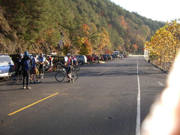 A group of cyclists preparing for a ride along a scenic road lined with parked cars. Fall foliage on the surrounding hills adds vibrant colors to the landscape. The sun casts a warm glow on the scene, creating a lively and inviting atmosphere for outdoor activities. Tanasi Trail System mountain bike trail.