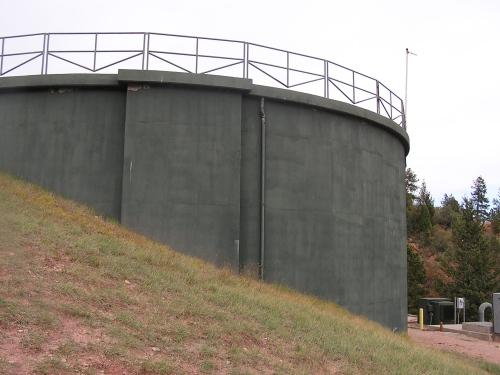 A green cylindrical water tank situated on a grassy hillside, with a metal railing at the top. Trees are visible in the background, indicating a natural setting. Blodgett mountain bike trail.