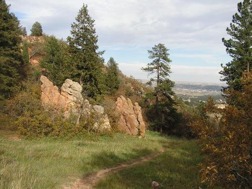 A rocky outcrop surrounded by lush green grass and trees, with a winding dirt path leading into the distance and a view of a valley below under a partly cloudy sky. Blodgett mountain bike trail.
