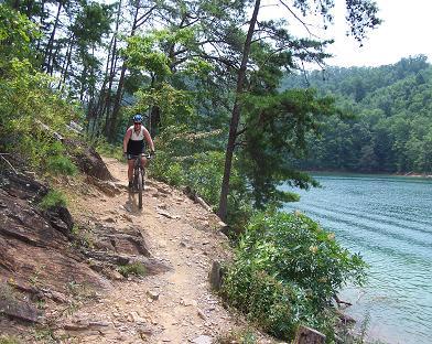 A person riding a mountain bike on a narrow trail beside a calm lake, surrounded by trees and hillside. The vibrant green foliage contrasts with the blue water, creating a scenic outdoor setting. Tsali Left Loop mountain bike trail.