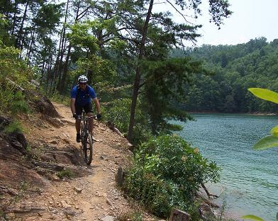 A cyclist riding a mountain bike along a rocky trail by a calm lake, surrounded by trees and greenery. Tsali Left Loop mountain bike trail.