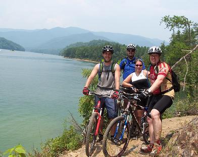 Four mountain bikers posing by a lake surrounded by green hills. They are wearing helmets and athletic clothing, with their bikes parked beside them. The scenic backdrop features a calm body of water and distant mountains under a bright blue sky. Tsali Left Loop mountain bike trail.