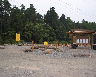 A gravel area with large rocks scattered around, featuring a wooden information kiosk with a roof and multiple signs. In the background, there are trees and a cloudy sky, indicating a natural outdoor setting. Two yellow signs are visible near the kiosk. USNWC mountain bike trail.