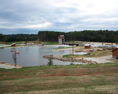A scenic view of an outdoor recreation area featuring a lake surrounded by grassy hills and wooded areas. In the background, there is a tall observation tower and several buildings, including a boathouse and additional facilities, with a cloudy sky overhead. USNWC mountain bike trail.