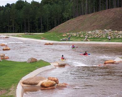 A scenic view of a river with several kayakers navigating the water. The riverbank features grassy areas and large rocks, with trees lining the background. Some people can be seen walking along the riverbank. USNWC mountain bike trail.
