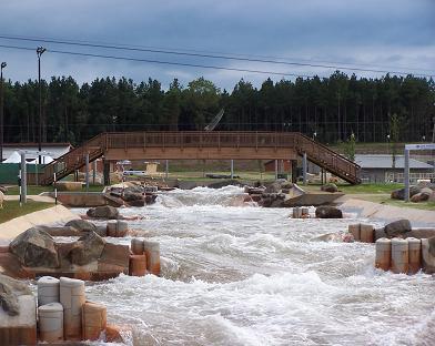 A view of a man-made whitewater rapids course with flowing water, surrounded by rocks and wooden structures. In the background, there is a bridge and trees, with a cloudy sky above. USNWC mountain bike trail.