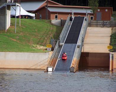 A kayak descends a ramp into a body of water, with a grassy area and buildings in the background. The scene shows a recreational area designed for water sports. USNWC mountain bike trail.