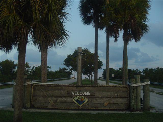 A wooden welcome sign at the entrance of a road lined with palm trees, leading into a scenic area. The sky is slightly overcast, suggesting early morning or late afternoon lighting. Oleta River State Park mountain bike trail.