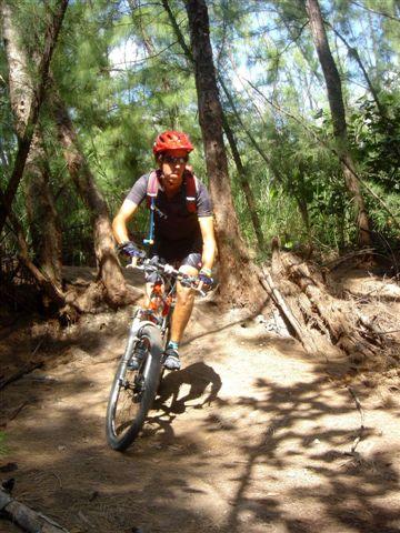 A person riding a mountain bike along a dirt trail in a wooded area, surrounded by trees. The rider is wearing a red helmet and a backpack, navigating the uneven terrain under dappled sunlight. Oleta River State Park mountain bike trail.