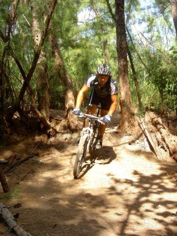 A person riding a mountain bike on a dirt trail surrounded by trees. The cyclist is wearing a helmet and a sporty outfit, navigating through a wooded area with sunlight filtering through the foliage. Oleta River State Park mountain bike trail.