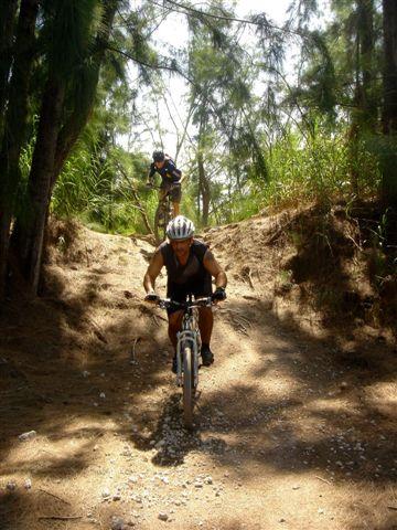 A person riding a mountain bike downhill on a sandy trail surrounded by trees, with another cyclist in the background navigating the same path. The scene is bright and captures an adventurous outdoor setting. Oleta River State Park mountain bike trail.