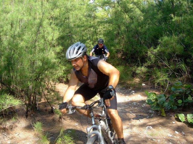 A mountain biker navigating a dirt trail surrounded by dense greenery, with another cyclist visible in the background. The biker in the foreground is wearing a helmet and athletic gear, focused on the path ahead. Oleta River State Park mountain bike trail.