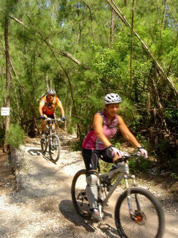 Two mountain bikers ride on a narrow trail surrounded by lush greenery. One cyclist, wearing a pink tank top and black pants, is in the foreground with an enthusiastic expression, while another cyclist in an orange and black outfit follows closely behind. Sunlight filters through the trees, illuminating the scene. Oleta River State Park mountain bike trail.