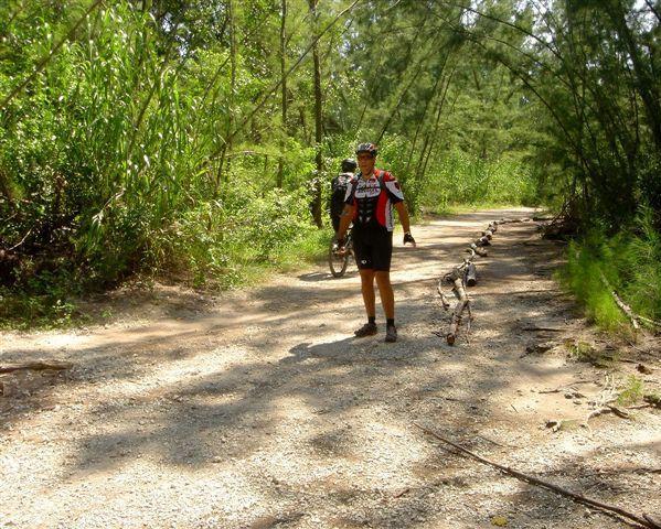 A cyclist standing on a gravel trail surrounded by lush greenery, with another cyclist in the background. The scene is illuminated by sunlight filtering through the trees, creating a serene outdoor atmosphere. Oleta River State Park mountain bike trail.