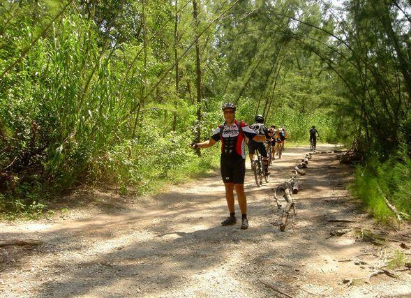 A group of cyclists rides on a gravel path through a lush, green forest. One cyclist stands in the foreground with arms outstretched, while others continue riding in the background. Sunlight filters through the trees, creating a vibrant, natural setting. Oleta River State Park mountain bike trail.