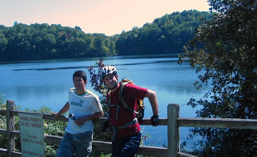 Two men stand beside a wooden fence overlooking a calm lake surrounded by trees in autumn colors. One man wears a white shirt with "Brasil" printed on it, while the other is dressed in a red shirt and a helmet, suggesting they have been biking. A sign nearby reads "PRIVATE PROPERTY DO NOT TRESPASS." The sky is clear and blue, creating a peaceful outdoor setting. Silver Comet Rail Trail mountain bike trail.