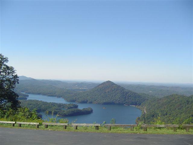A scenic view of a mountain and lake surrounded by lush greenery, with rolling hills and clear blue skies in the background. The image captures the tranquility of the natural landscape, highlighting the reflective water and varied terrain. Tanasi Trail System mountain bike trail.
