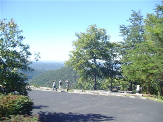 A scenic view of a mountain landscape under a clear blue sky, featuring several people standing and taking photos. In the foreground, there are trees and a paved area, with distant mountains visible beyond. Tanasi Trail System mountain bike trail.