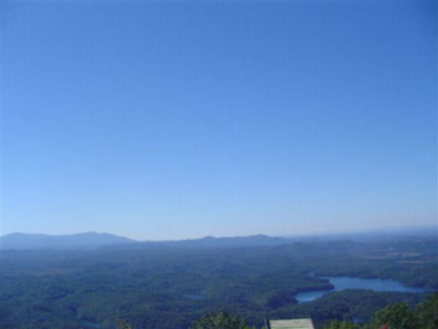 A panoramic view of a mountainous landscape under a clear blue sky, featuring lush green hills and a winding lake in the foreground. Tanasi Trail System mountain bike trail.