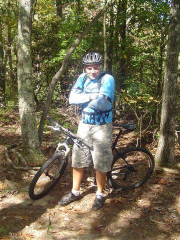 A person in a blue cycling jersey and helmet stands confidently with arms crossed beside a mountain bike, surrounded by a wooded area with trees and foliage. Tanasi Trail System mountain bike trail.
